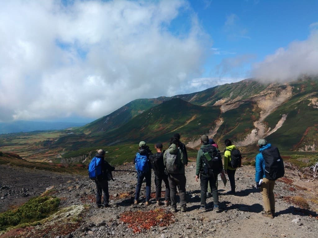 山岳の登山道で撮影クルーが機材を携え、谷と山並みを見渡すロケ風景